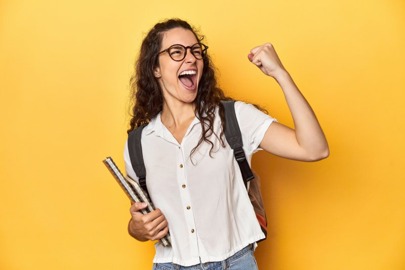 Young woman wearing glasses holding books in one hand and fist pumping the air with the other