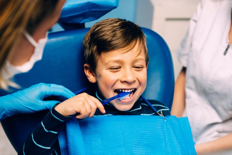 Young boy smiling in dental chair as dentist instructs him how to brush his teeth